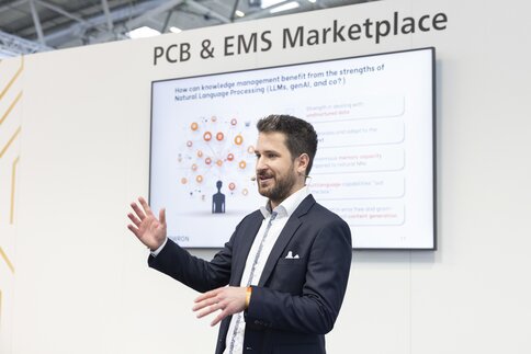 A man with short hair and wearing a suit is holding a microphone and giving a presentation in front of a screen that says 