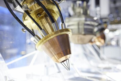 Nahaufnahme von Industriemaschinen mit einer Art Düse aus Metall und Schläuchen die damit verbunden sind. Im Hintergrund sind unscharf weitere Maschinen sichtbar.