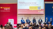 Six participants in the presentation are sitting in a semi-circle on the stage in Hall A1 of Air Cargo Europe and chatting. The title of the presentation, a QR code and the Transport Logistic logo can be seen in the background. The audience is blurred in the foreground.