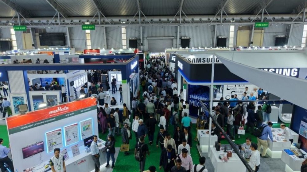 A crowded technical exhibition hall with booths, banners, and lots of people walking around and chatting.