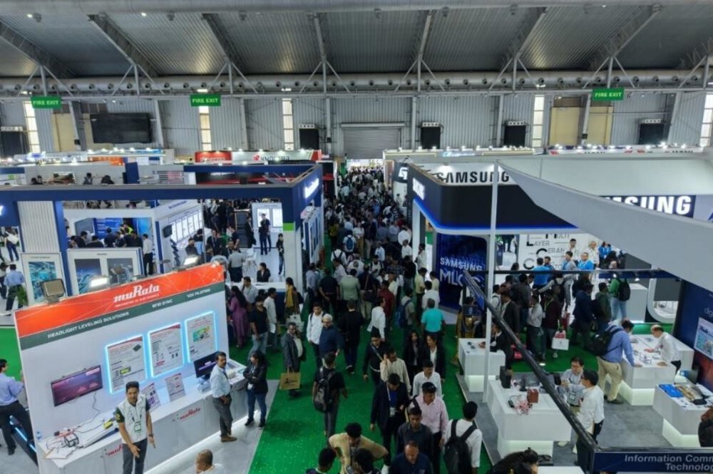 A crowded technical exhibition hall with booths, banners, and lots of people walking around and chatting.