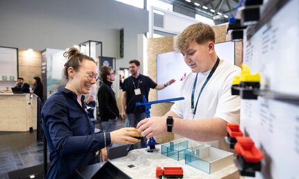 Am Messestand sind eine junge Frau und ein junger Mann im Gespräch. Auf dem Tisch ist ein kleiner Kran und ein kleiner Lastwagen zu sehen.