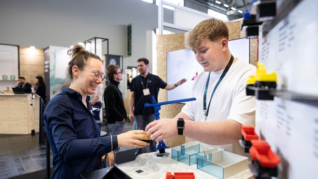 Am Messestand sind eine junge Frau und ein junger Mann im Gespräch. Auf dem Tisch ist ein kleiner Kran und ein kleiner Lastwagen zu sehen.