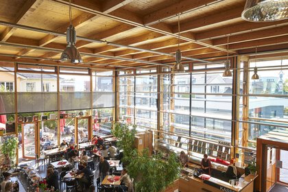 Geräumiges Café mit großen Fenstern, Holzdecke, Zimmerpflanzen und Leuten, die an Tischen sitzen.