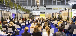 Ein mit Stellwänden abgetrennter Bereich der BAU, in dem ein sitzendes Publikum einem Sprecher lauscht, der einen Vortrag auf der Bühne hält.