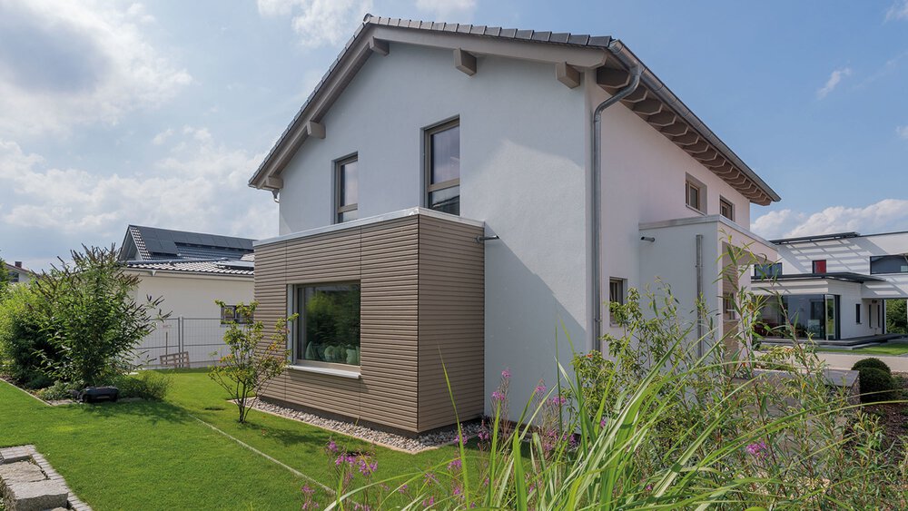 Ein modernes weißes Haus mit flachem Anbau, umgeben von grünem Rasen und Pflanzen, unter einem teilweise bewölkten Himmel.