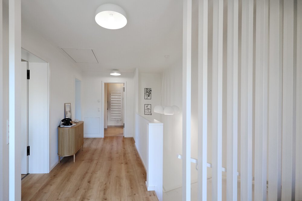 Heller Flur mit Holzboden, weißen Wänden und vertikalen weißen Lamellen neben einem Treppenabgang.