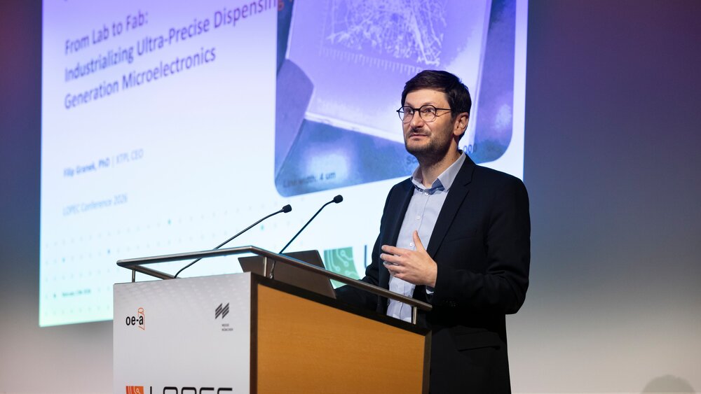 A man in a suit and glasses is standing at a lectern at LOPEC in front of a screen showing a presentation.