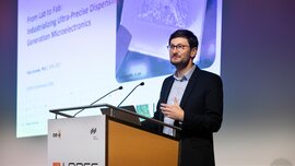 A man in a suit and glasses is standing at a lectern at LOPEC in front of a screen showing a presentation.