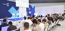 Eine Frau spricht auf einer Bühne mit hoher blauer Rückwand vor einem sitzenden Publikum auf einer Messe.