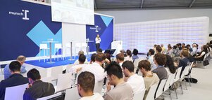 A woman speaks on a stage with a tall blue backdrop in front of a seated audience at a trade fair.