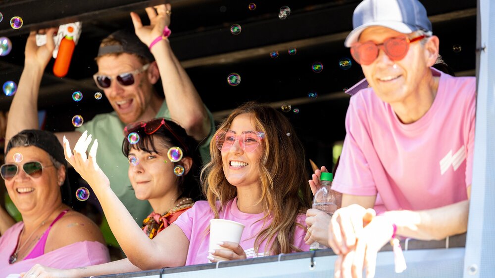 Five people in pink shirts, smiling and playing with soap bubbles at an outdoor event.