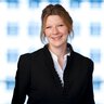 Eine Frau im schwarzen Blazer, mit weißer Bluse posiert lächelnd.