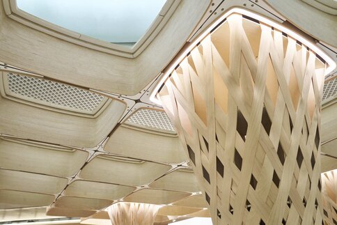 A woven wooden column supports a ceiling made of geometric wooden panels with skylight openings.