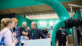 Technical discussion at a green trade fair stand in front of a giant turquoise agitator propeller for water treatment.