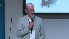 Ein Mann im hellgrauen Blazer spricht auf der Bühne in ein Mikrofon, hinter ihm eine Leinwand mit einem Bild.