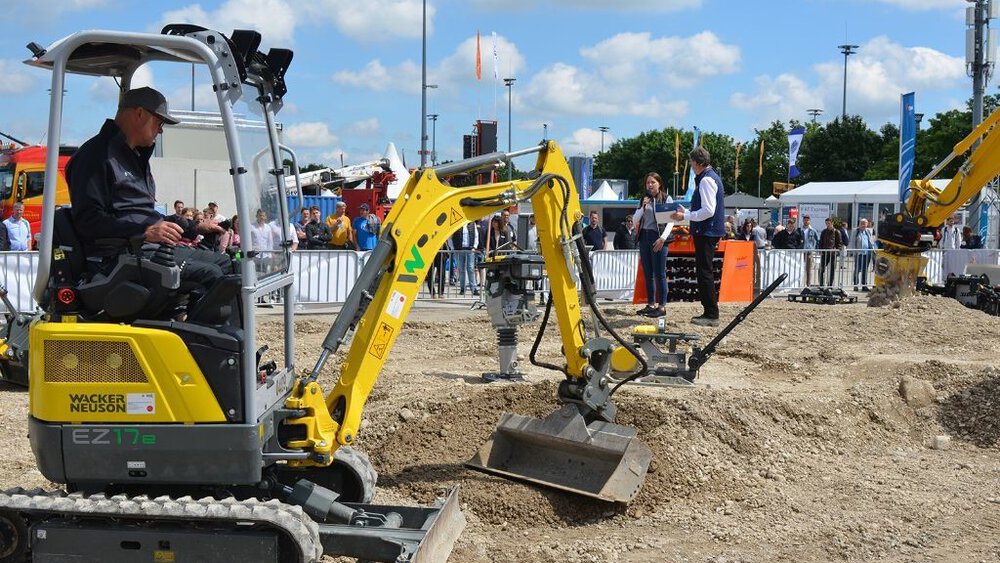 A small yellow excavator is digging in an open-air demonstration area, people and tents can be seen in the background.