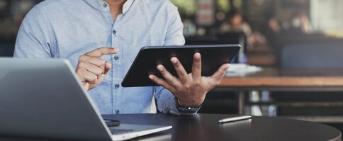 Der Oberkörper eines Mannes, der ein Tablet in den Händen hält. Neben ihm liegen ein Laptop und ein Smartphone.