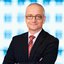 Ein Mann mit dünner schwarzer Brille, einem dunklen Anzug und roter Krawatte posiert mit Lächeln und verschränkten Armen.