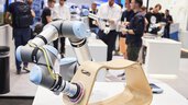 A robotic arm at an exhibition stand polishes a complex, rounded wooden stool mounted on an exhibition table.