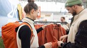 Two people are standing at an outdoor clothing booth, looking at an item of clothing together. In the background, more jackets and textiles are displayed on clothes racks.