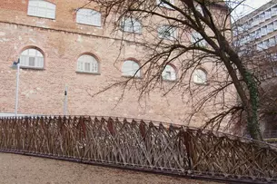 Wooden lattice bridge with an intricate design next to a brick building and leafless trees.