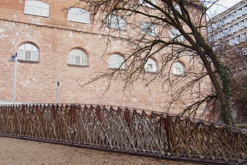 Holzgitterbrücke mit kompliziertem Design neben einem Backsteingebäude und blattlosen Bäumen.