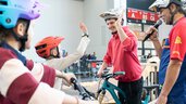 A young man wearing a bicycle helmet gives a child on a bicycle a high five during a participatory activity in the exhibition hall. Next to him stands a presenter with a microphone, and in the background you can see barriers and spectators.