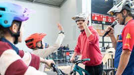 A young man wearing a bicycle helmet gives a child on a bicycle a high five during a participatory activity in the exhibition hall. Next to him stands a presenter with a microphone, and in the background you can see barriers and spectators.