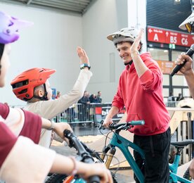 A young man wearing a bicycle helmet gives a child on a bicycle a high five during a participatory activity in the exhibition hall. Next to him stands a presenter with a microphone, and in the background you can see barriers and spectators.