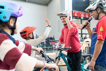 Ein junger Mann mit Fahrradhelm gibt einem Kind auf einem Fahrrad ein High-Five während einer Mitmachaktion in der Messehalle. Daneben steht ein Moderator mit Mikrofon, im Hintergrund sind Absperrgitter und Zuschauer zu sehen.