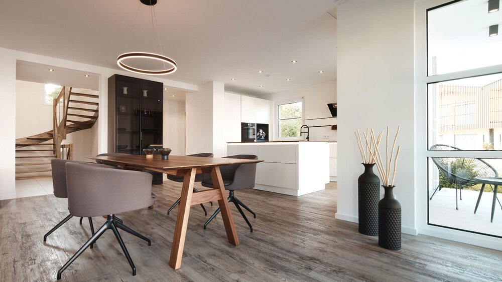 Moderner Essbereich mit Holztisch, grauen Stühlen und offener Küche; große Fenster und Treppenhaus sichtbar.