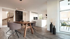 Moderner Essbereich mit Holztisch, grauen Stühlen und offener Küche; große Fenster und Treppenhaus sichtbar.