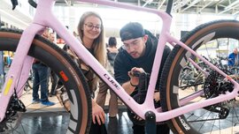 Two people are standing at a bicycle trade fair booth, examining a racing bike on display on a presentation stand. The exhibition hall with other visitors can be seen in the background.