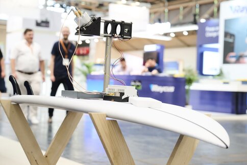 Technische Vorrichtung mit Kabeln und Sensoren auf einem weißen Surfboard als Träger, präsentiert auf einer Messe als Teil moderner Lasersysteme.