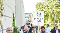 Menschen folgen im Atrium zwischen den Messehallen einer Person mit dem Schild „IFAT Munich Solution Tours“.