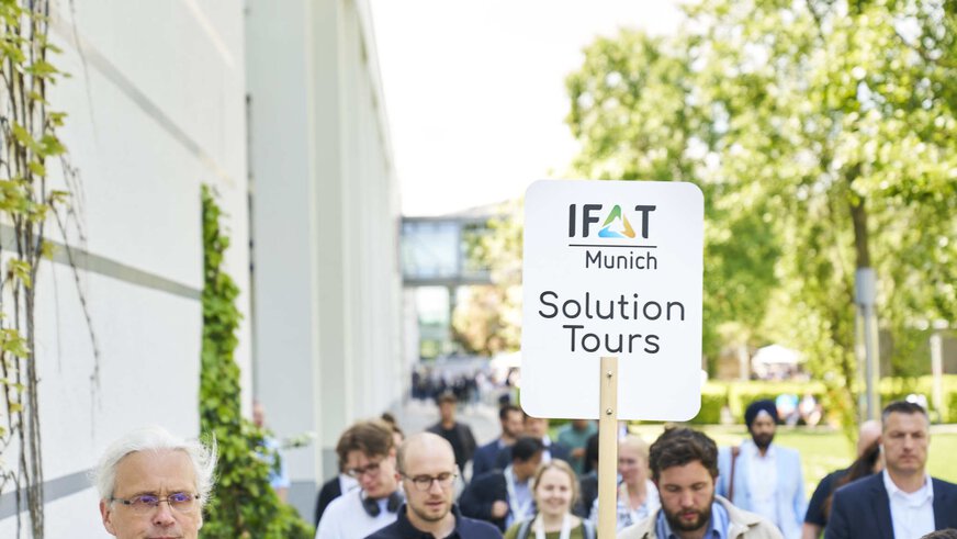 Menschen folgen im Atrium zwischen den Messehallen einer Person mit dem Schild „IFAT Munich Solution Tours“.