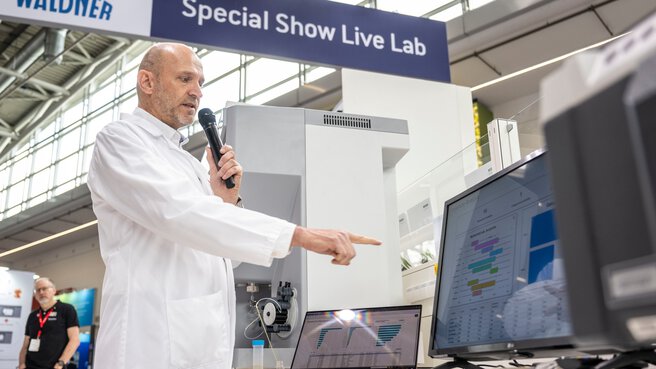A scientist in a white lab coat presents analysis results on a screen at the “Special Show Live Lab” at analytica.