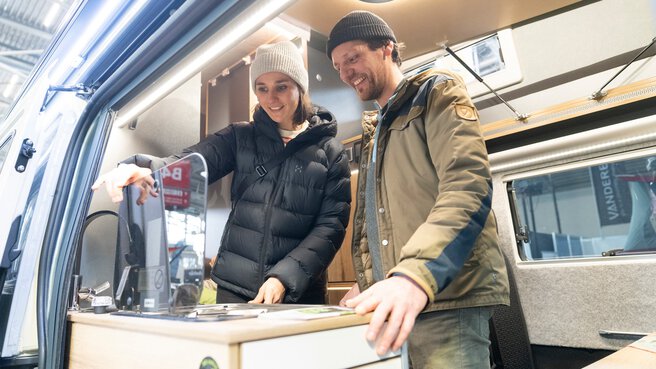 Two people are standing in a converted camper van, looking at the kitchenette together. In the background, you can see cabinets, lighting, and other details of the vehicle's interior.