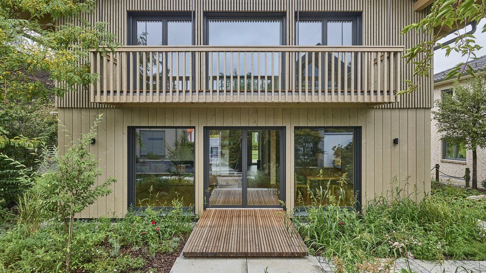 Modernes zweistöckiges Holzhaus mit Balkon, umgeben von Grün und einem Holzsteg, der zum Eingang führt.