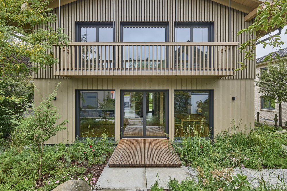 Modernes zweistöckiges Holzhaus mit Balkon, umgeben von Grün und einem Holzsteg, der zum Eingang führt.