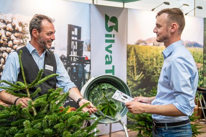Zwei Männer im Gespräch an einem Messestand, einer zeigt Tannenzweig, der andere hält Informationsmaterial in der Hand.