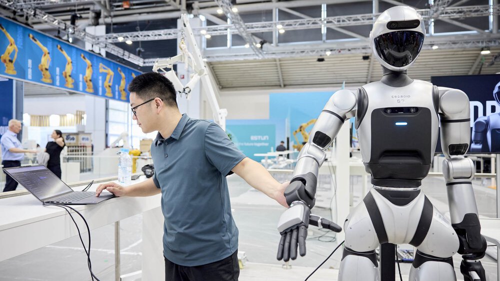 Ein Mann steht neben einem weiß-schwarzen humanoiden Roboter und betrachtet etwas auf einem Laptop. Im Hintergrund ist eine Messehalle mit weiteren Ständen zu sehen.