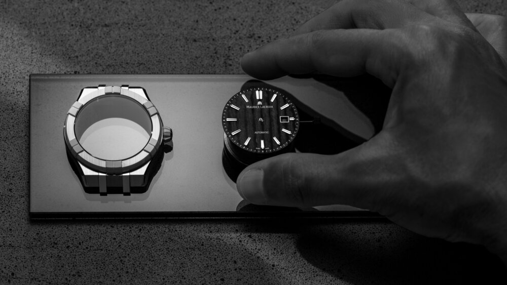 Black-and-white close-up of a hand reaching for the face of a Maurice Lacroix wristwatch. The watch case lies next to it.