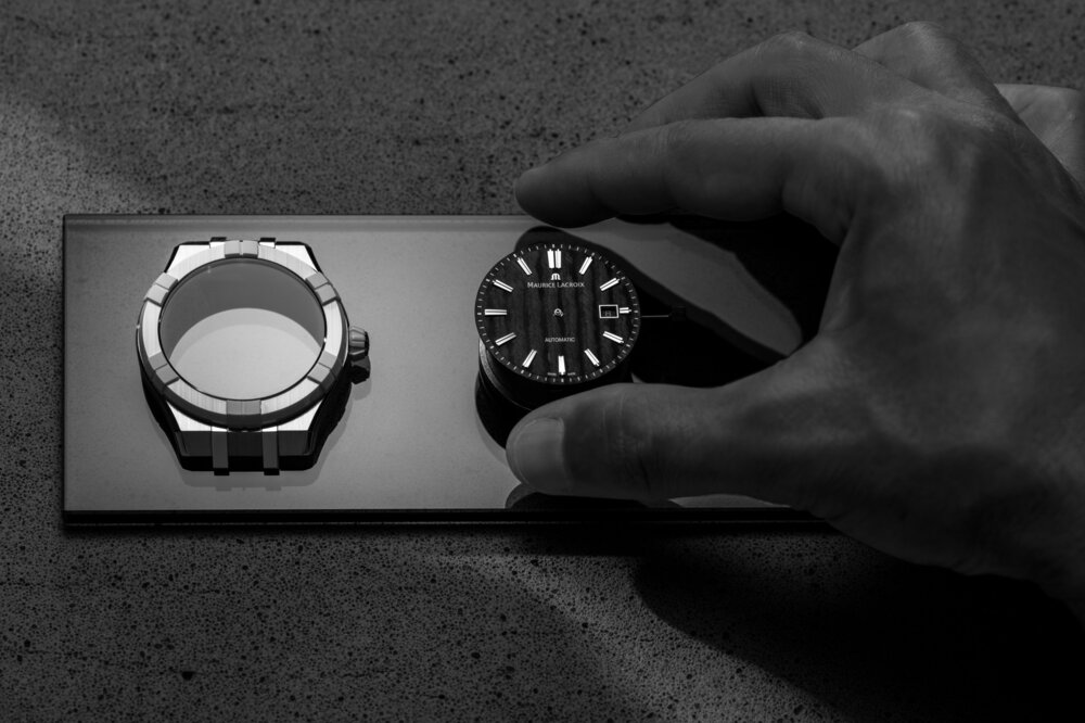 Schwarz-weiße Nahaufnahme einer Hand, die nach dem Ziffernblatt einer „Maurice Lacroix“ Armbanduhr greift. Daneben liegt das Gehäuse der Uhr.